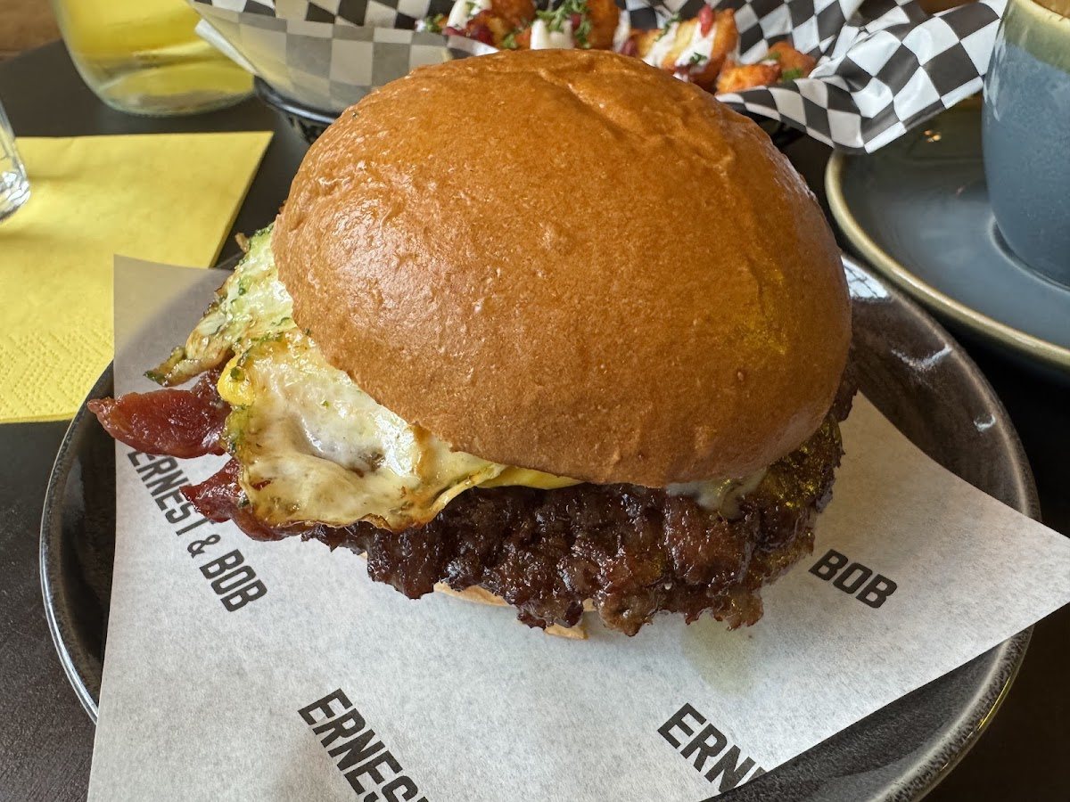 A smash burger on printed Ernest & Bob paper: a glossy brioche bun over crisp bacon, a fried egg, melted cheese and a deeply caramelised smashed patty.