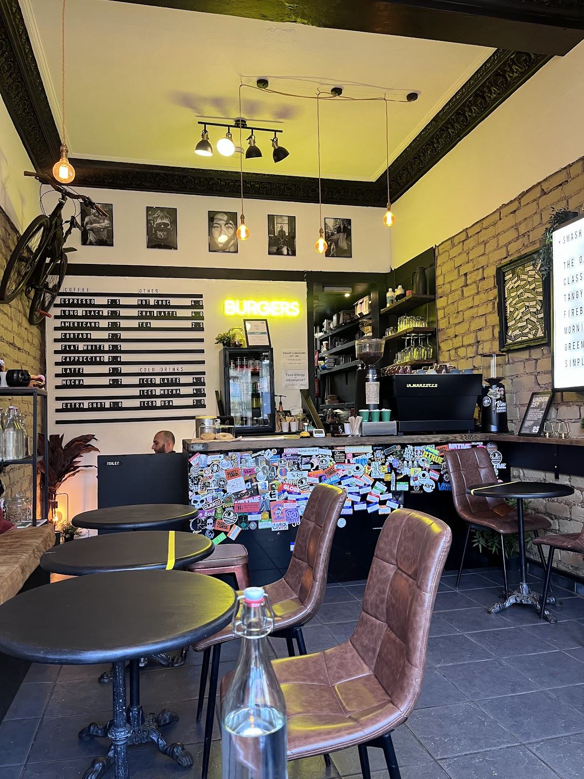 The counter: a coffee letterboard on the wall, a yellow neon BURGERS sign, a bicycle hung on the wall, and black-and-white portraits under ornate cornicing.
