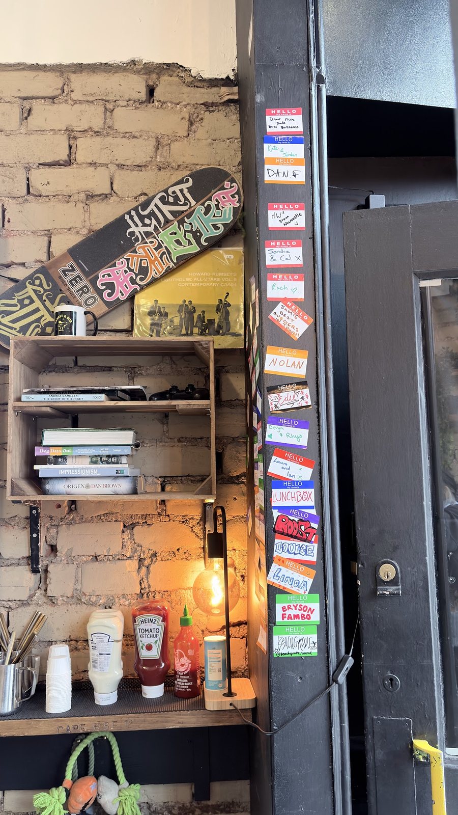 By the door: a column of colourful 'Hello my name is' stickers up the black frame, a skateboard deck, a crate shelf of books, and ketchup and sriracha bottles under a lamp.