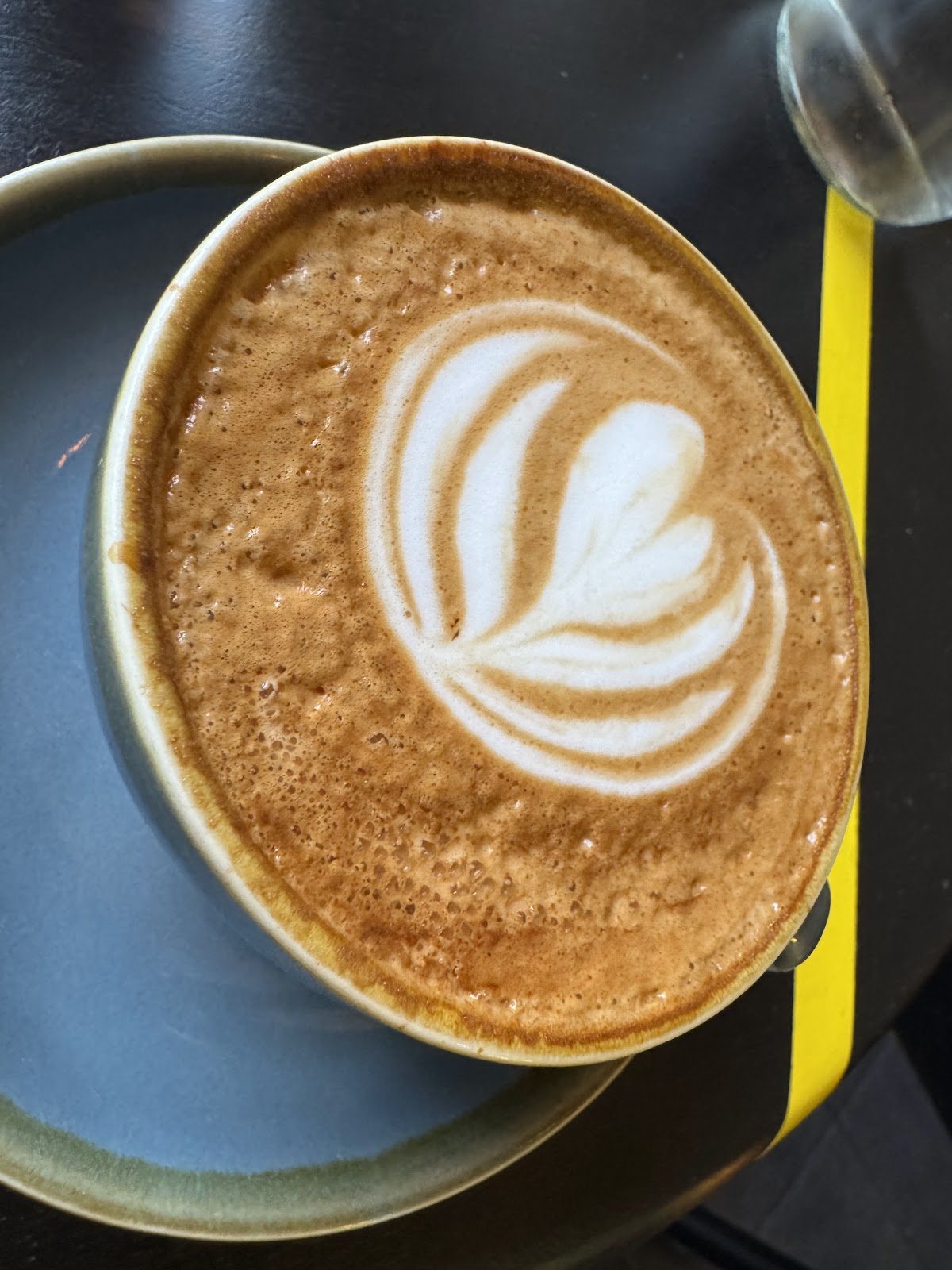 A flat white with a tulip in latte art, in a slate-blue cup on a black table with a yellow stripe.